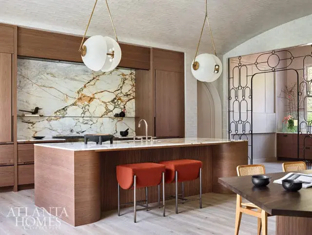 Modern kitchen with a large marble slab backsplash, wood cabinetry, and a waterfall island with orange stools. Two globe pendant lights hang above the island.