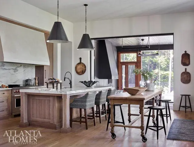Warm wood kitchen with marble island, gray upholstered stools, and large black pendant lights. A wooden table with rolling casters sits in front of a large window overlooking trees.
