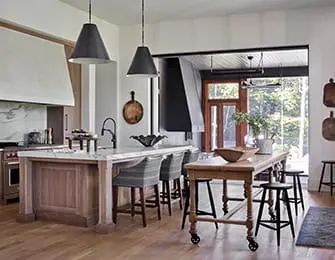 Spacious kitchen with a large island topped with white marble, dark wood cabinetry, and pendant lights. A wooden table with stools sits nearby, with a large open doorway leading outside.