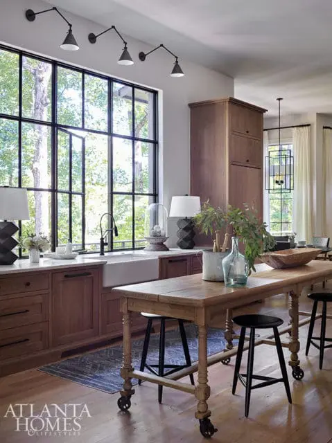 Kitchen with warm wood cabinetry, a large farmhouse sink, and a long wooden island with black stools. A wall of windows overlooks a wooded area.