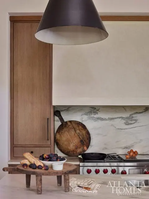 Kitchen with wood cabinetry, marble backsplash, stainless steel range, and a large black pendant light. A wooden cutting board with figs and bread sits on the counter.