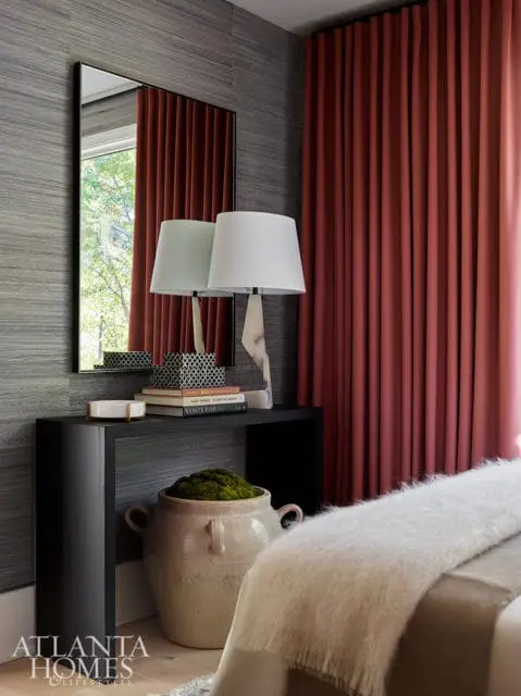 Bedroom with gray textured wallpaper, rust-colored curtains, a black console table with books and lamps, and a large ceramic pot filled with moss.