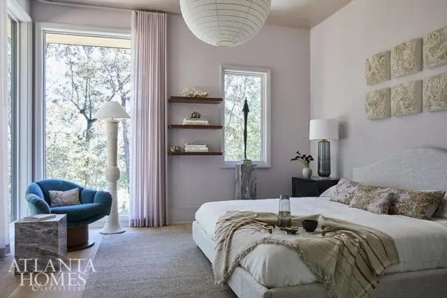 Bedroom with a large bed, blue armchair, and floor-to-ceiling windows overlooking trees. Walls are painted a soft lavender, with decorative art above the bed.
