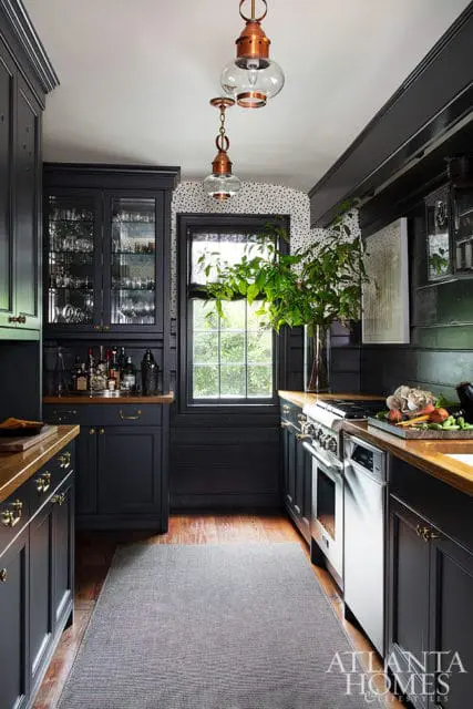 Dark gray kitchen with glass-front cabinetry, copper pendant lights, a stainless steel range, and a window with a large vase of greenery.