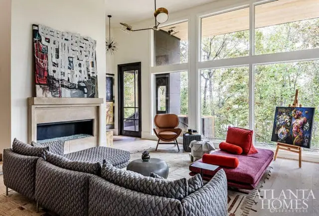 Living room with a stone fireplace, abstract art, gray quilted sofas, a red chaise lounge, and large windows overlooking trees.