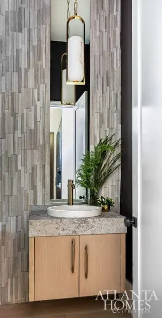 Powder room with a textured gray stone tile wall, a light wood vanity with brass hardware, a stone countertop, and a white vessel sink. A modern brass pendant light hangs above.