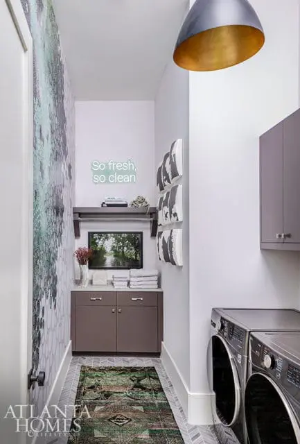 Laundry room with gray cabinets, stacked towels, a television displaying a nature scene, and a neon sign that reads 'So fresh, so clean.'