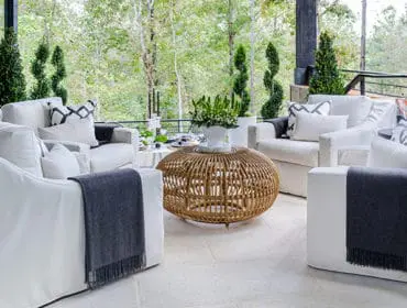 Covered patio with white slipcovered sofas and armchairs, a round rattan coffee table, and a lush green forest backdrop.