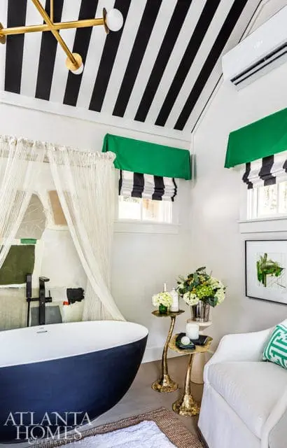 Modern bathroom with a navy freestanding bathtub, black and white striped ceiling, green and black striped Roman shades, and abstract art above the tub.