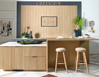 Modern kitchen island with fluted wood cabinetry, marble countertop, and two bar stools. Above the island, a wall features the word "CREEKSIDE" in gold lettering.