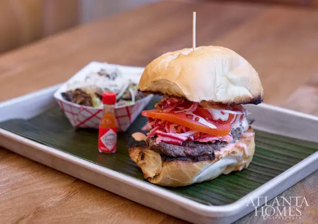 Close-up of a gourmet burger with tomato, slaw, and sauce, served with a side of fries and a mini Tabasco bottle on a metal tray lined with a banana leaf.