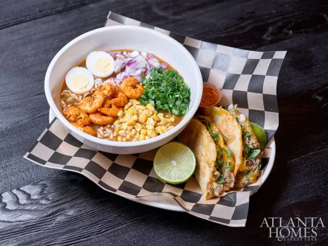 Bowl of ramen with shrimp, corn, egg, and red onion, served with three tacos and lime wedges on a checkered paper liner.