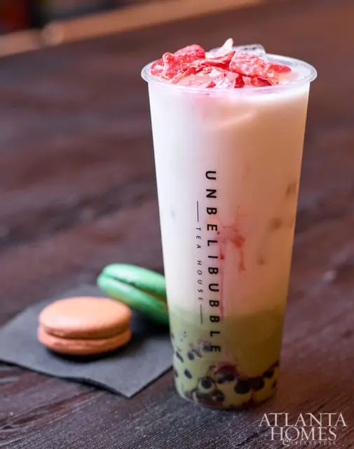 Tall plastic cup with layered pink and green bubble tea, topped with crushed ice and strawberry pieces. Two macarons, one orange and one green, sit on a dark napkin to the left.