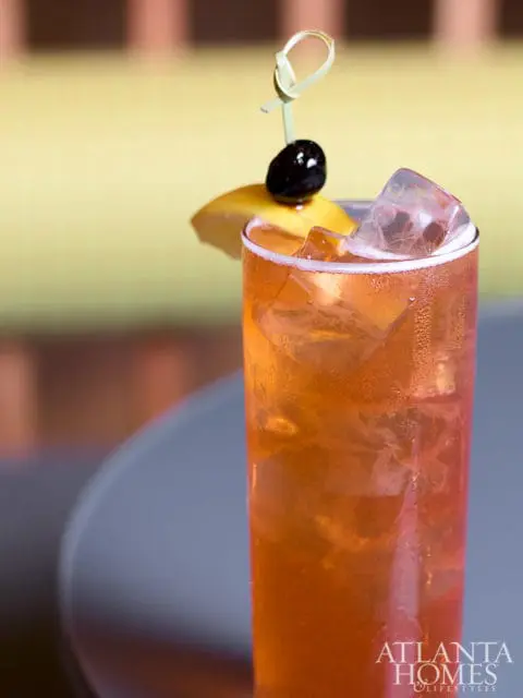 Tall glass filled with amber-colored iced drink, garnished with a lemon slice, cherry, and a decorative pick. Atlanta Homes & Lifestyles logo in the lower right corner.