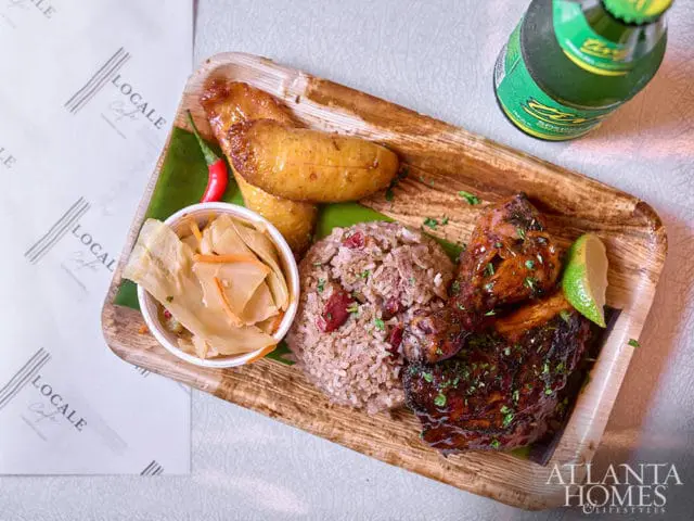 Plate of jerk chicken with rice and peas, fried plantains, pickled cabbage, and a lime wedge, served on a banana leaf with a bottle of sparkling beverage.