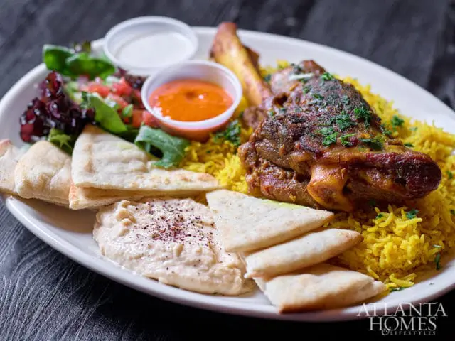 Plate of food with lamb shank, yellow rice, hummus, pita bread, salad, and dipping sauces, with Atlanta Homes & Lifestyles logo in the corner.