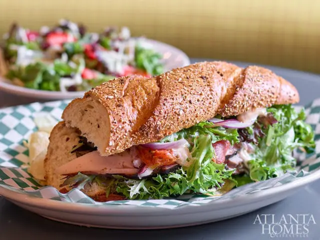 Close-up of a large sandwich on a seeded roll with turkey, lettuce, tomato, and onion, served with potato chips and a side salad in the background.