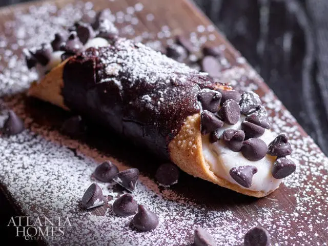 Cannoli filled with cream, dipped in dark chocolate, and topped with chocolate chips and powdered sugar, on a wooden board with scattered chocolate chips.