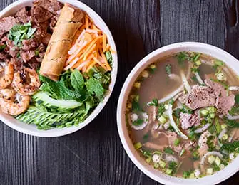 Two bowls of Vietnamese food: one with noodles, beef, and herbs; the other a rice bowl with spring roll, shrimp, beef, and vegetables.