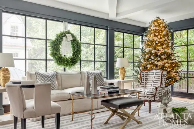 Living room with a large Christmas tree, a green wreath hanging on the window, a white sofa, and patterned armchairs.