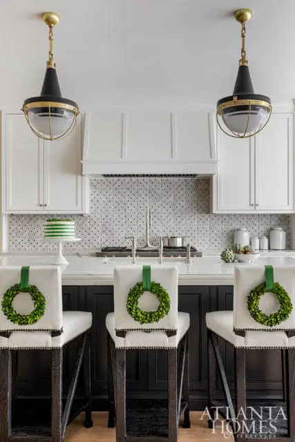 White kitchen with black island, white bar stools adorned with boxwood wreaths, and industrial-style pendant lights.