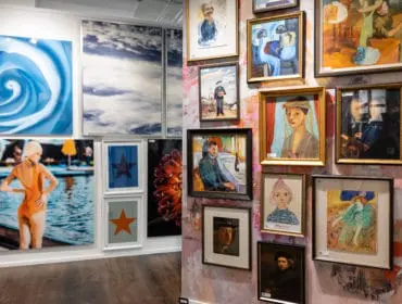 Gallery wall with diverse artwork including a blue rose print, sky photographs, a woman in an orange swimsuit, abstract stars, a floral close-up, and various framed paintings of people and scenes.