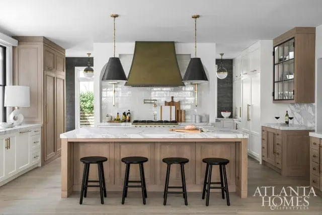Luxury kitchen with a large island featuring a marble countertop and wood base, surrounded by black stools. White subway tile backsplash, brass accents, and a prominent brass range hood.