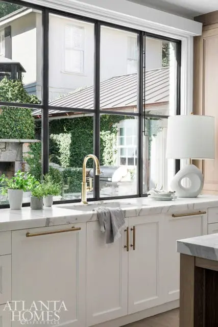 White kitchen with marble countertops, brass faucet, and white cabinetry with brass hardware, overlooking a lush green backyard through a large black-paned window.