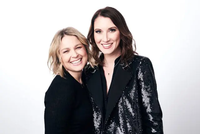 Two women smiling at the camera, one with blonde hair wearing a black sweater, the other with brown hair wearing a black sequined jacket.