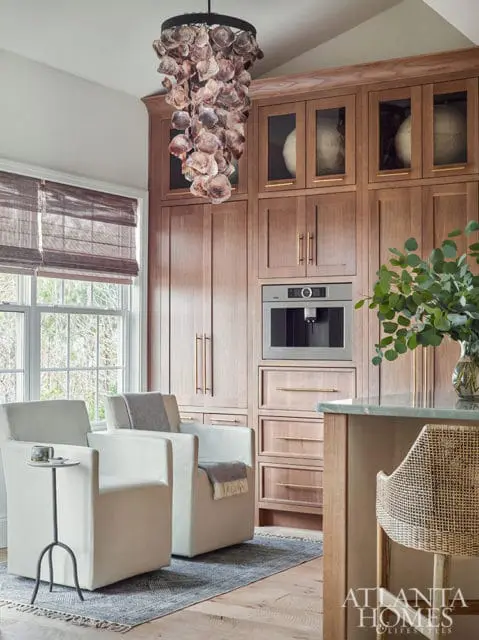 Modern kitchen with built-in coffee maker, light wood cabinetry, and two white armchairs. A unique shell chandelier hangs above.