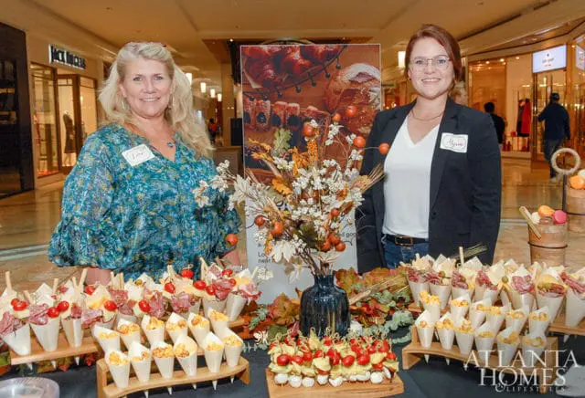 Two women stand behind tables laden with appetizers. The woman on the left wears a teal patterned blouse and the woman on the right wears a black blazer over a white v-neck shirt. A floral arrangement with fall foliage sits between them.