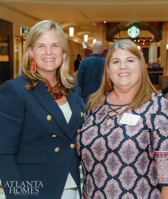 Two women smiling, one in a navy blazer with gold buttons and large orange necklace, the other in a patterned dress with a name tag that reads "Lisa Suttles".
