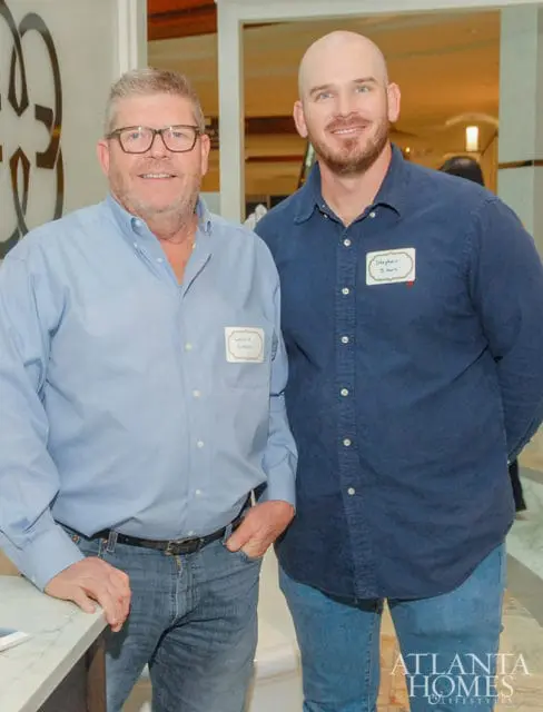 Two men, Lonnie Simon and Stephen Simon, wearing blue shirts and jeans. Lonnie Simon wears glasses and has his hand in his pocket. Stephen Simon has a bald head and a goatee.