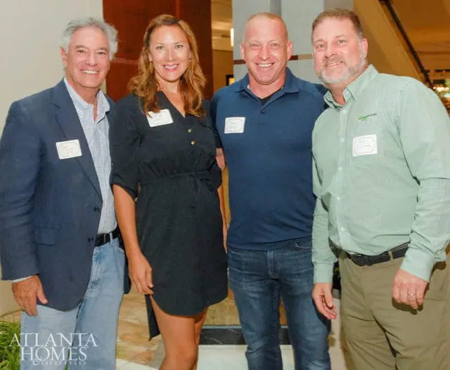 Four people smiling for a photo, two men and two women. The man on the far left wears a navy blazer and jeans. The woman next to him wears a black dress. The man in the middle wears a navy polo shirt and jeans. The man on the far right wears a green checkered shirt and khaki pants.