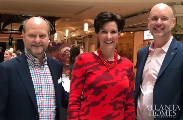 Three people smiling at the camera, two men in suits and a woman in a red patterned sweater and coral necklace. The Atlanta Homes & Lifestyles logo is visible in the lower right corner.