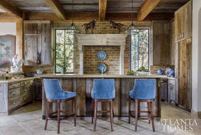 Rustic kitchen with reclaimed wood cabinetry and island, brick backsplash, stone countertops, and blue velvet bar stools.