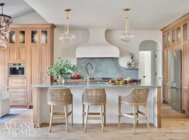 Light wood kitchen with a large island topped with a marble countertop, woven bar stools, and a pale green marble backsplash behind the range.