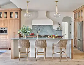 Light wood kitchen with a large island topped with a light-colored countertop, three woven bar stools, and a pale green backsplash.