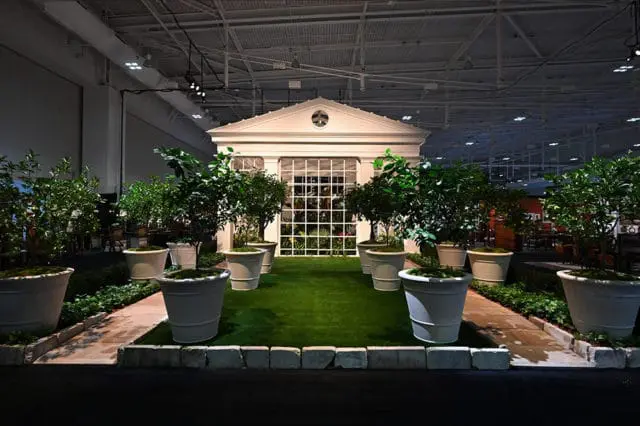 Indoor garden display with a white classical facade structure, large potted trees, and artificial turf