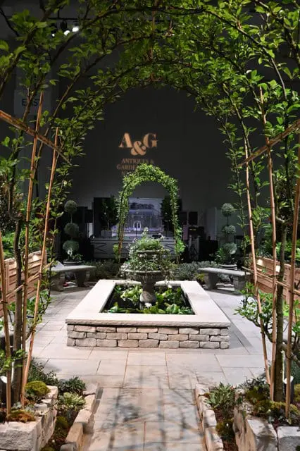 Stone pathway leading to a formal garden with a tiered fountain in a rectangular pond, flanked by benches and arched trellises covered in greenery.