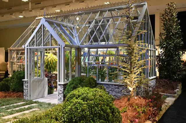 Glass greenhouse with white trim and stone base, featuring an open door and lush greenery inside and out.