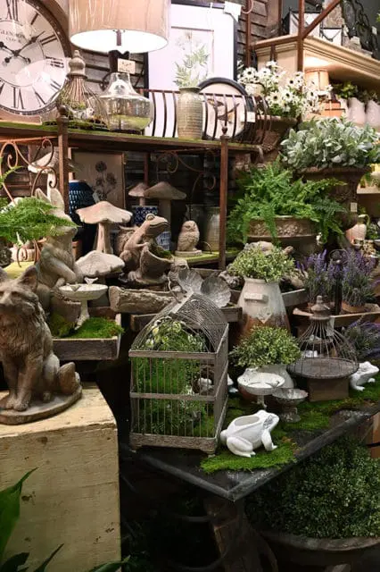 Display of garden-themed decor including a large clock, ceramic frogs, mushroom statues, birdcages, and potted plants.