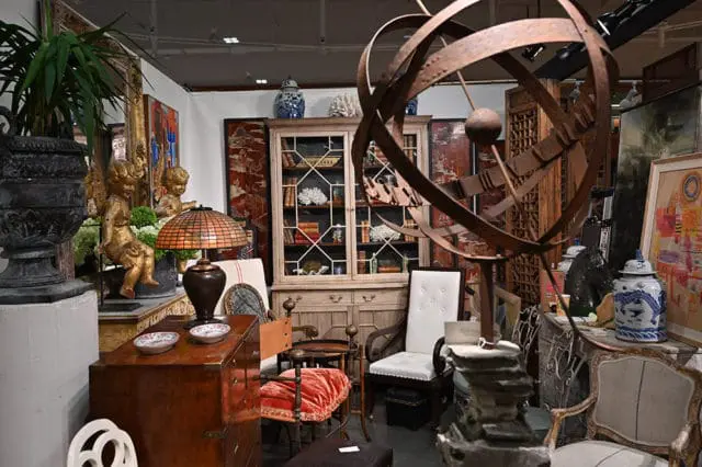 Antique shop interior with a large metal armillary sphere, a glass-fronted cabinet filled with books, and various decorative items including statues and porcelain vases.