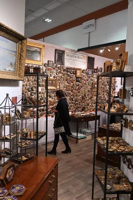 Woman browsing a display of hexagonal and square decorative items, many with gold accents, on a wall and shelves. A sign reads 'MUSEUM BEES'.