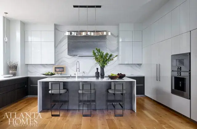 Modern kitchen with white high-gloss cabinetry, dark lower cabinets, a gray waterfall island with bar stools, and a stainless steel range hood over a marble backsplash.