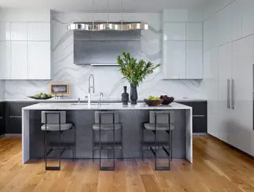 Modern kitchen with white and dark wood cabinetry, a gray waterfall island with bar stools, marble backsplash, and stainless steel appliances.