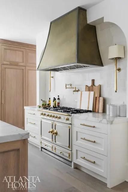 White kitchen with a large brass range hood, white subway tile backsplash, marble countertops, and light wood cabinetry.