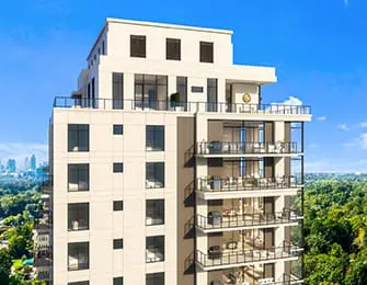 Modern high-rise building with cream-colored facade, glass balconies, and dark-framed windows, surrounded by lush green trees under a clear blue sky.