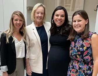 Four women smiling at the camera indoors. The woman on the far left wears a black blazer over a white top. The second woman from the left wears a cream blazer over a navy dress. The third woman from the left wears a black dress. The woman on the far right wears a colorful floral dress.