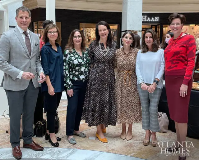 Seven people posing for a photo indoors, with a man in a grey suit on the left and six women in dresses and slacks. The Atlanta Homes & Lifestyles logo is in the bottom right corner.